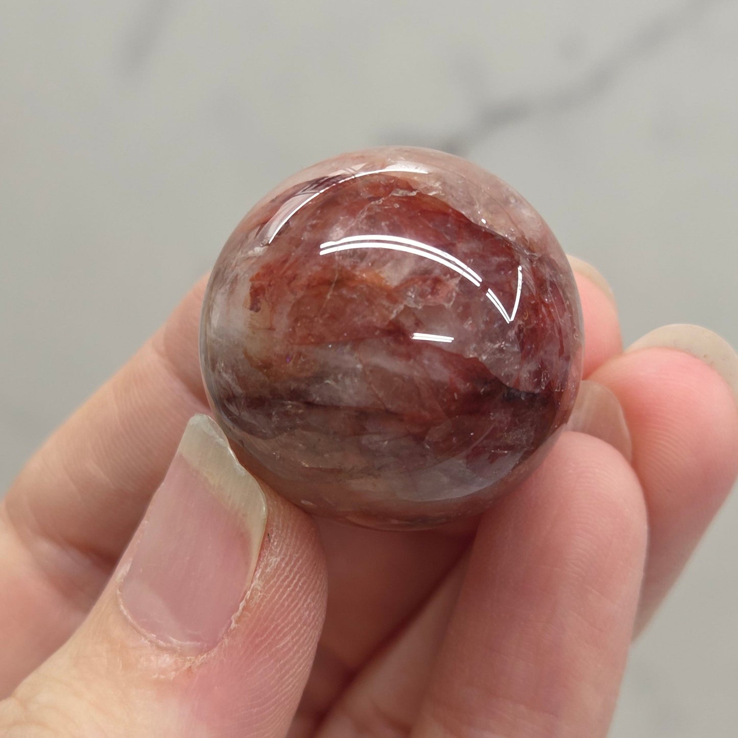 A hand holding a polished round gemstone with a mottled reddish-brown and white pattern, set against a light gray marble background.