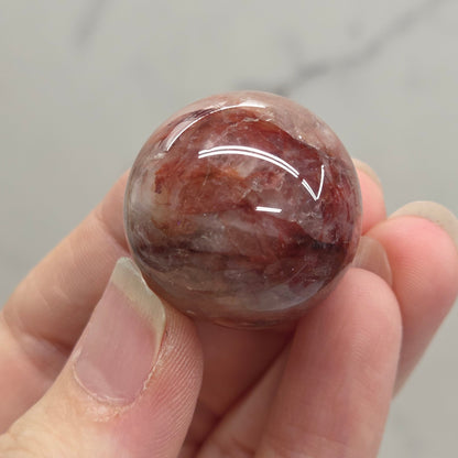 A hand holding a polished round gemstone with a mottled reddish-brown and white pattern, set against a light gray marble background.