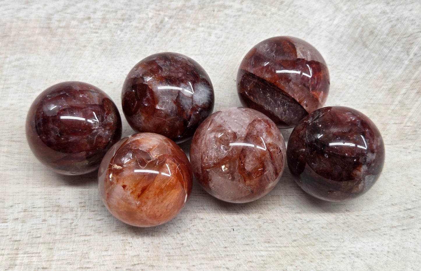 Five polished round gemstone beads in earthy brown, burgundy, and smoky tones, arranged on a light beige textured fabric background.