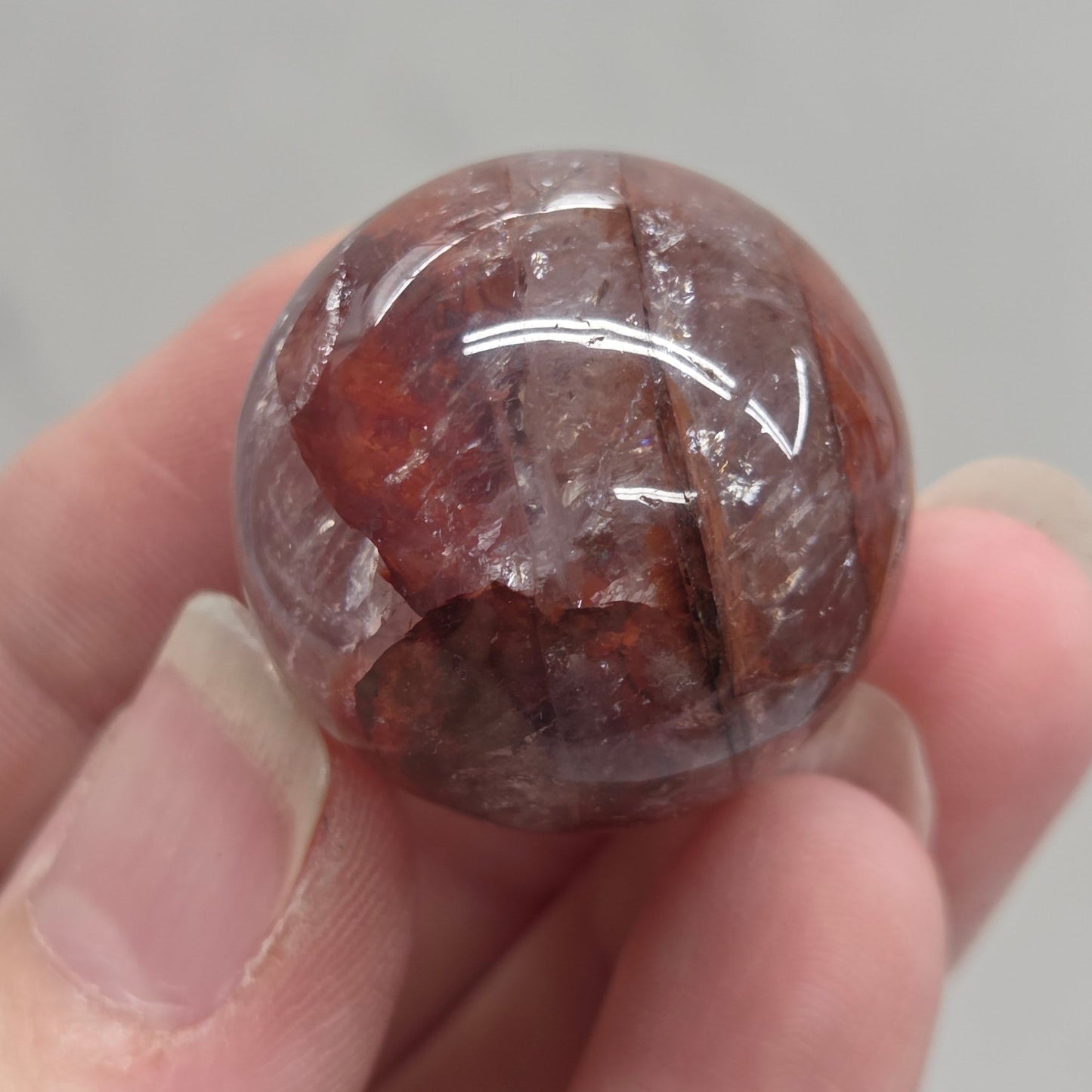 A hand holding a polished round gemstone with a reddish-brown and clear crystalline structure against a light gray marble background.