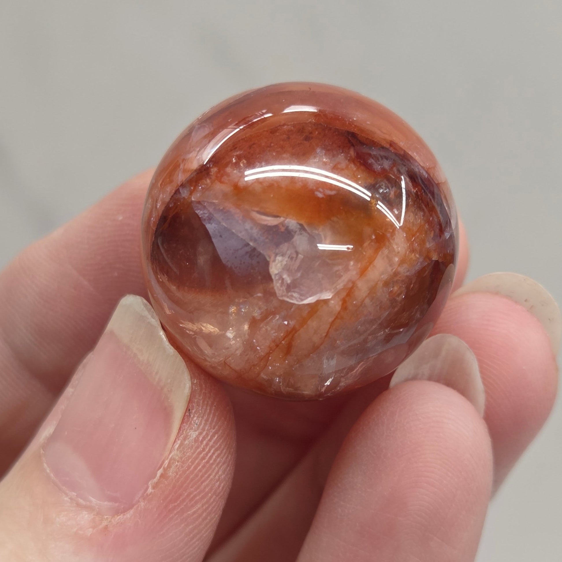 A hand holding a polished round gemstone with swirling amber-brown and translucent qualities against a light gray marble-like background.