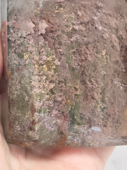 A close-up of a hand holding a rectangular translucent stone or crystal with a mottled earthy pattern in brown, green, and gray tones.