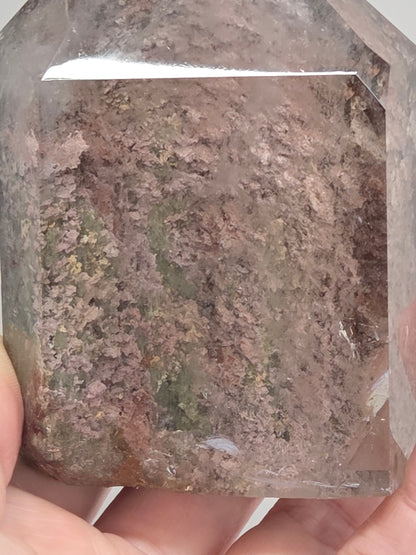 A hand holding a large, faceted clear crystal or glass stone with dense, textured internal inclusions in shades of brown, gray, and muted green.