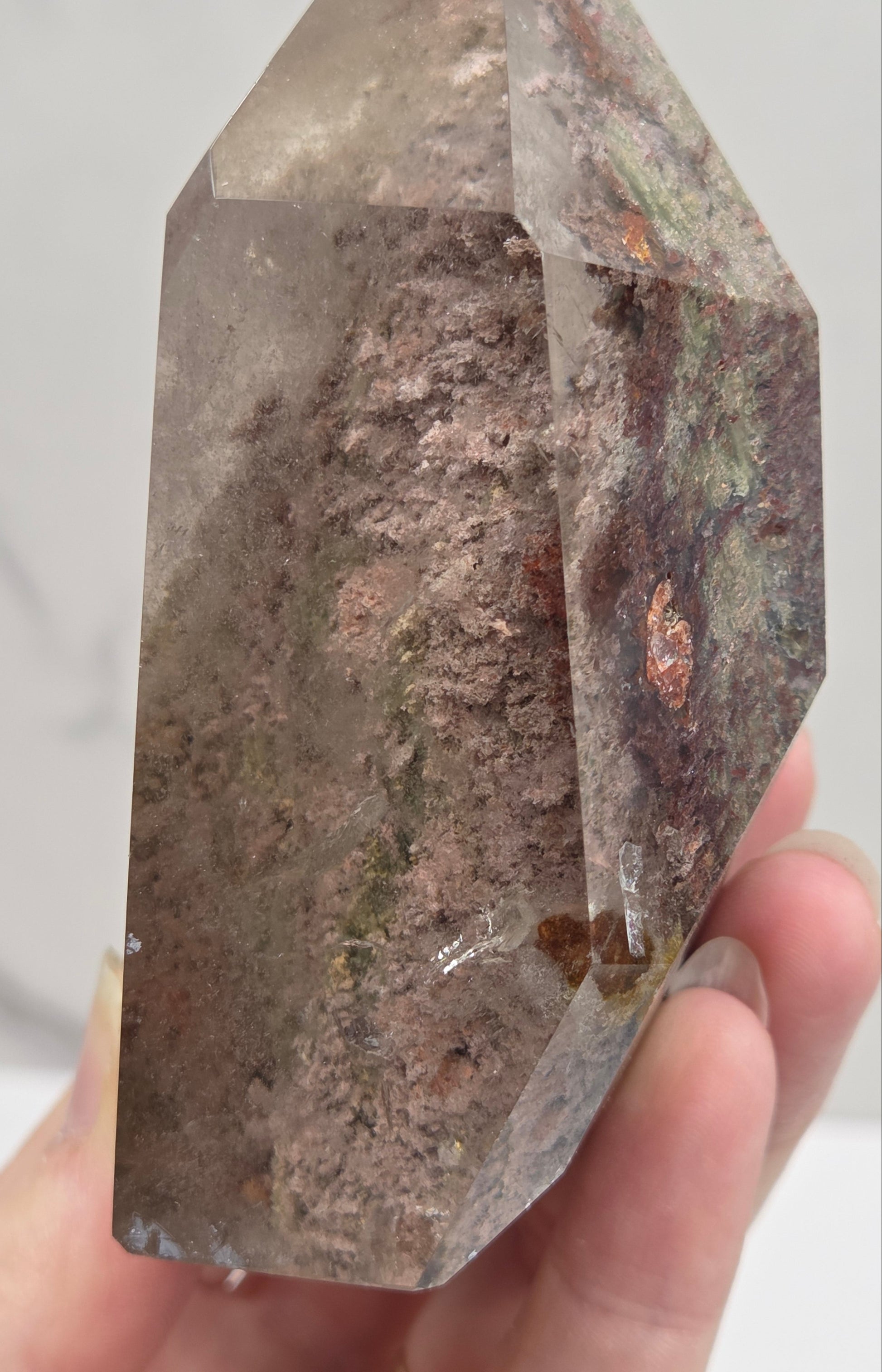 A hand holding a raw, faceted crystal point with a smoky gray and brown textured interior and subtle copper and green inclusions, set against a light gray marble background.