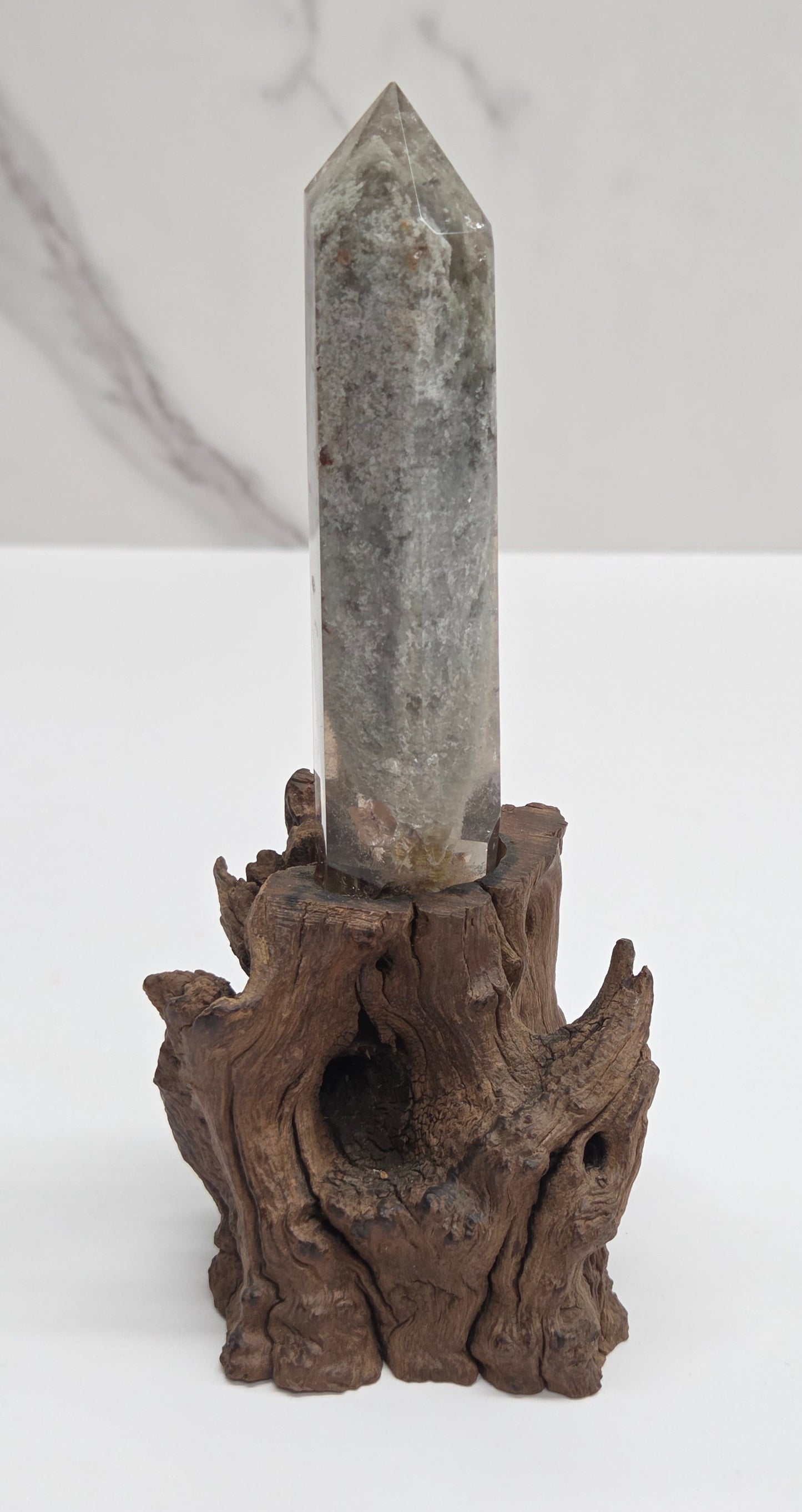 A gray crystal point spear tip mounted on a piece of dark brown driftwood, displayed against a split white and light gray marble background.