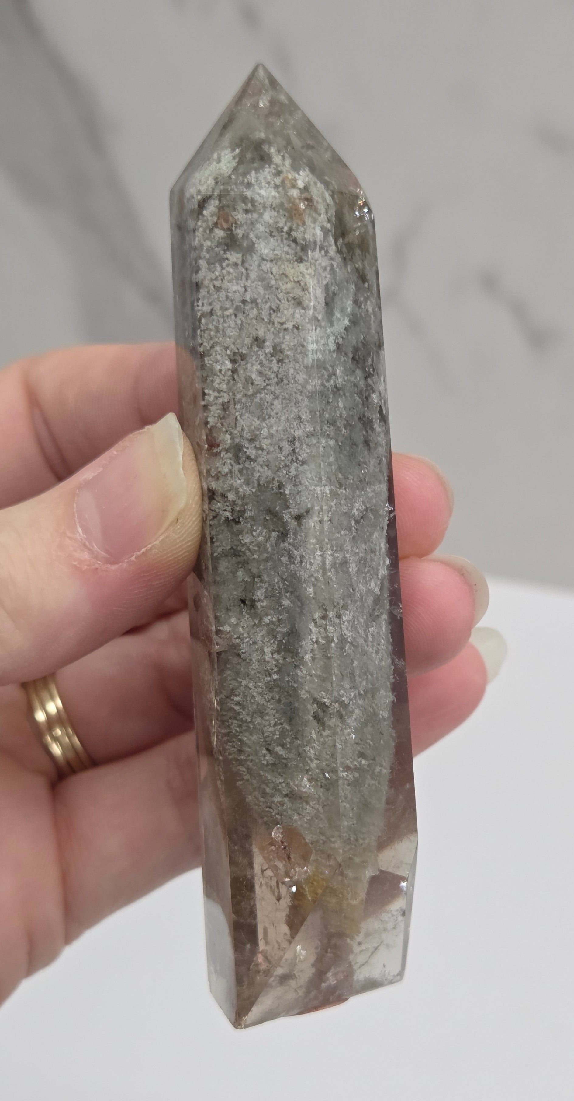 A person's hand holding a raw, pointed crystal tower with a gray and white marbled texture, set against a light gray marble-patterned background.