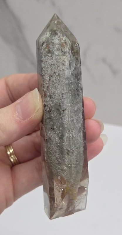 A person's hand holding a raw, pointed crystal tower with a gray and white marbled texture, set against a light gray marble-patterned background.