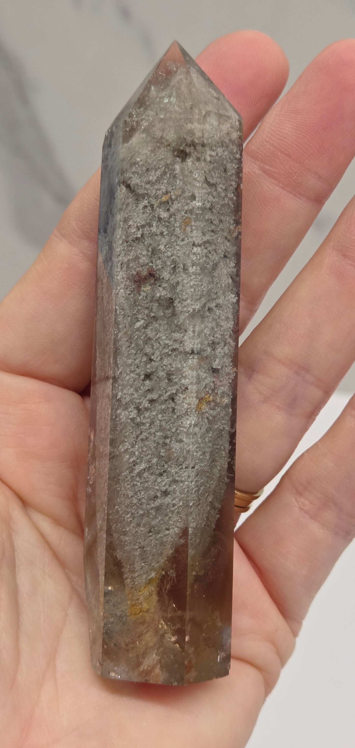 A person's hand holding a polished natural quartz crystal point with a gray and brown speckled texture.