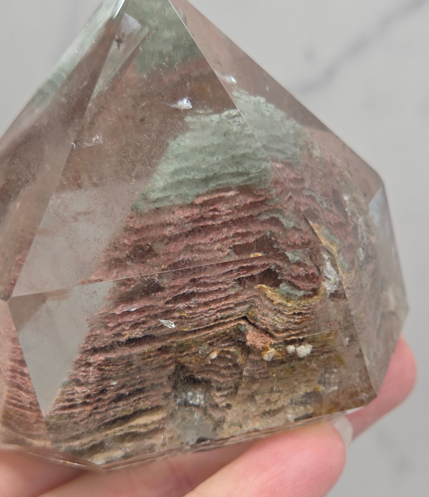 A faceted clear crystal point, likely quartz, held in a hand to show its internal brown and reddish-brown layered inclusions, against a light gray marble background.