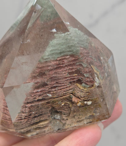 A faceted clear crystal point, likely quartz, held in a hand to show its internal brown and reddish-brown layered inclusions, against a light gray marble background.