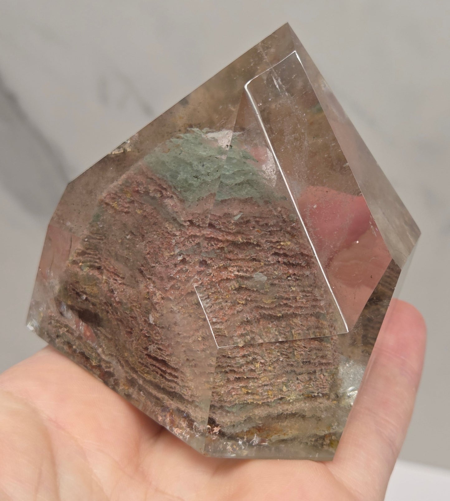 A hand holding a clear faceted crystal or gemstone with a textured, earthy brown and green inlay or resin-like center, shown against a light marble-like background.