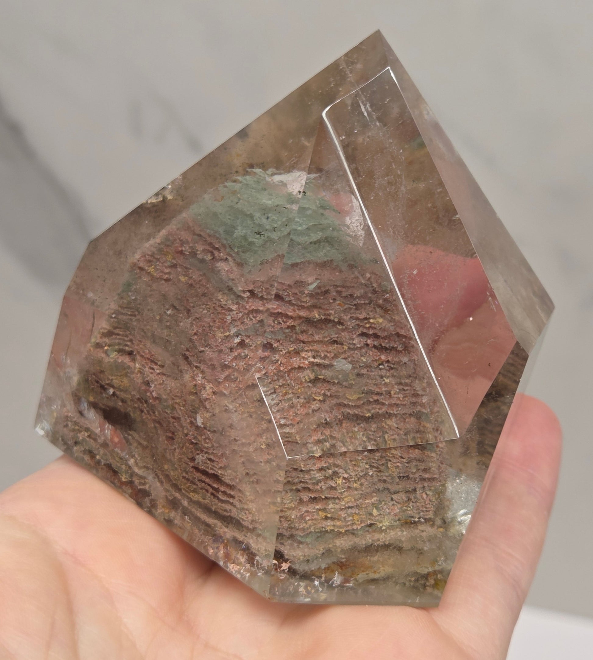 A hand holding a clear faceted crystal or gemstone with a textured, earthy brown and green inlay or resin-like center, shown against a light marble-like background.