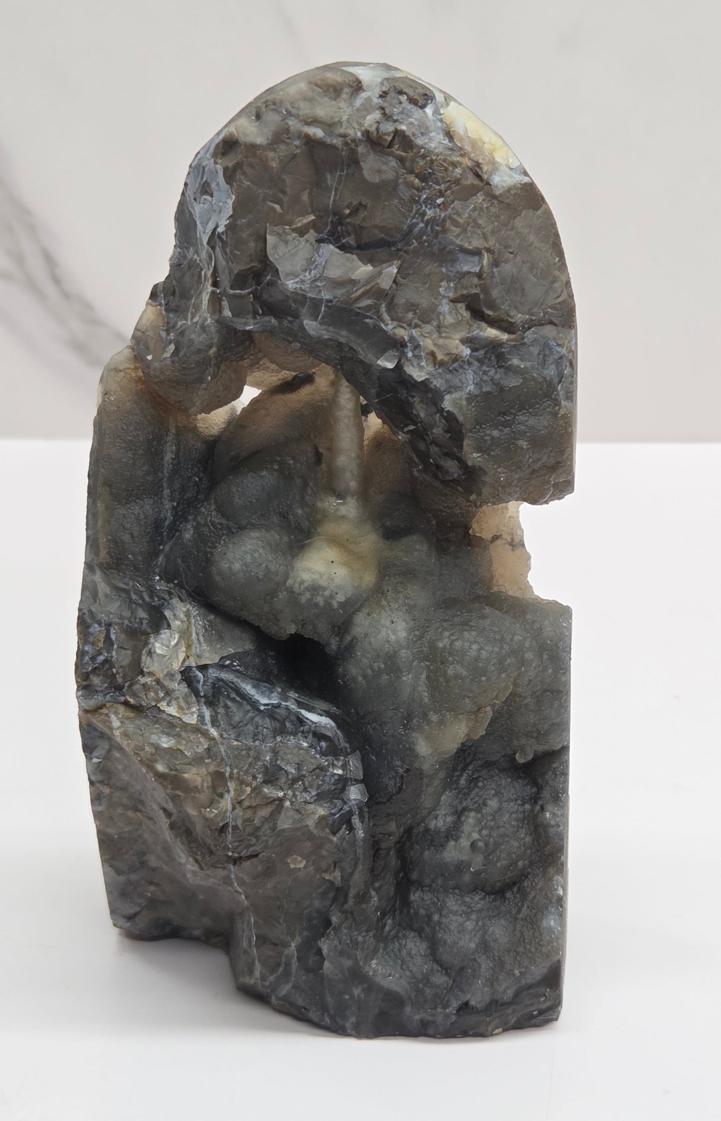 A volcano agate free form with subtle beige and blue-gray veining, standing on a white surface against a light gray marble-patterned background.