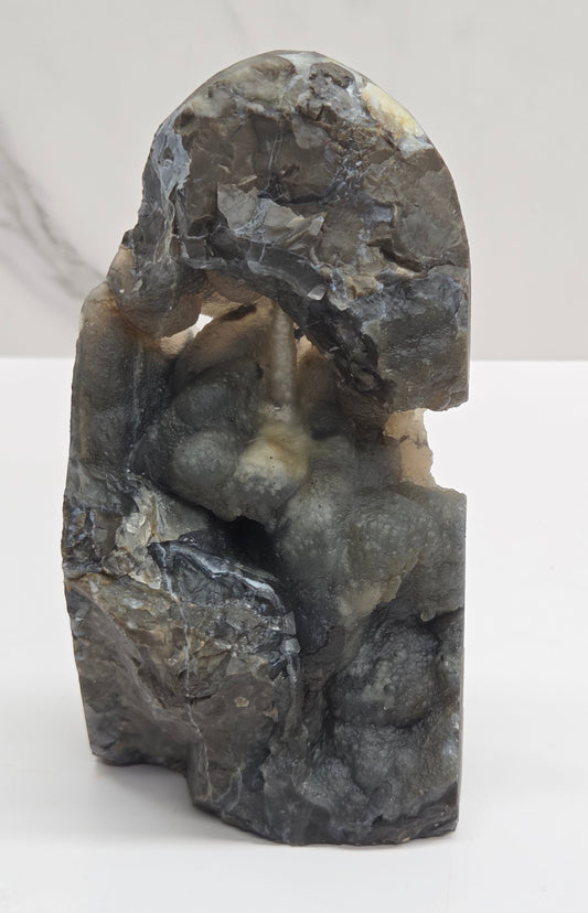 A volcano agate free form with subtle beige and blue-gray veining, standing on a white surface against a light gray marble-patterned background.
