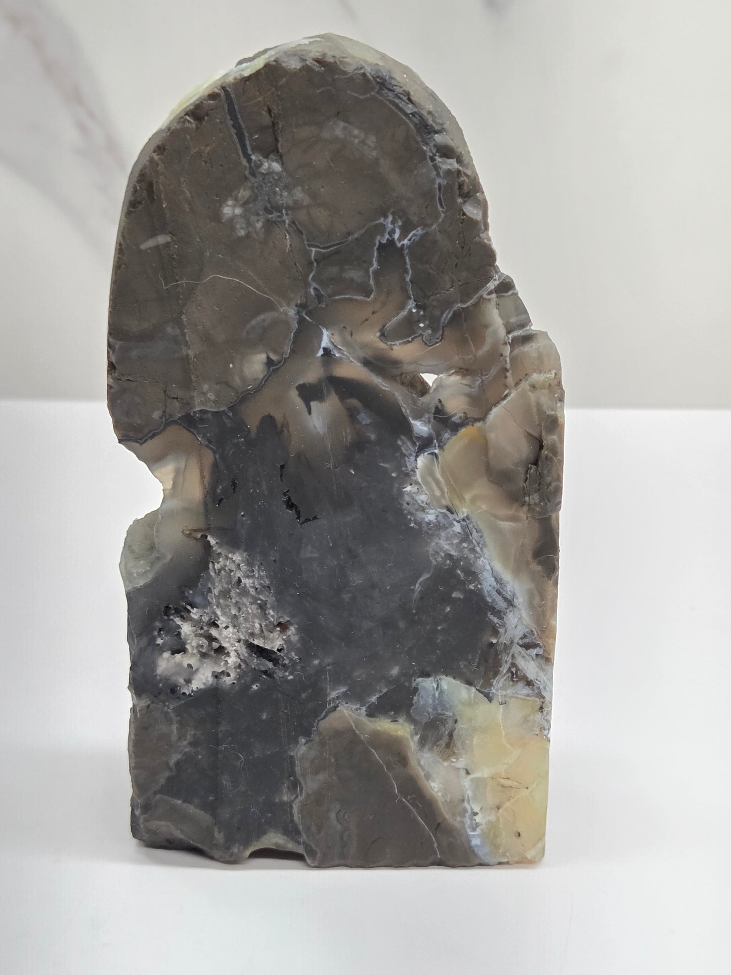 A raw, polished stone carving piece with organic, irregular shapes, featuring natural patterns of gray, black, beige, and white, standing upright on a white surface against a light gray background.