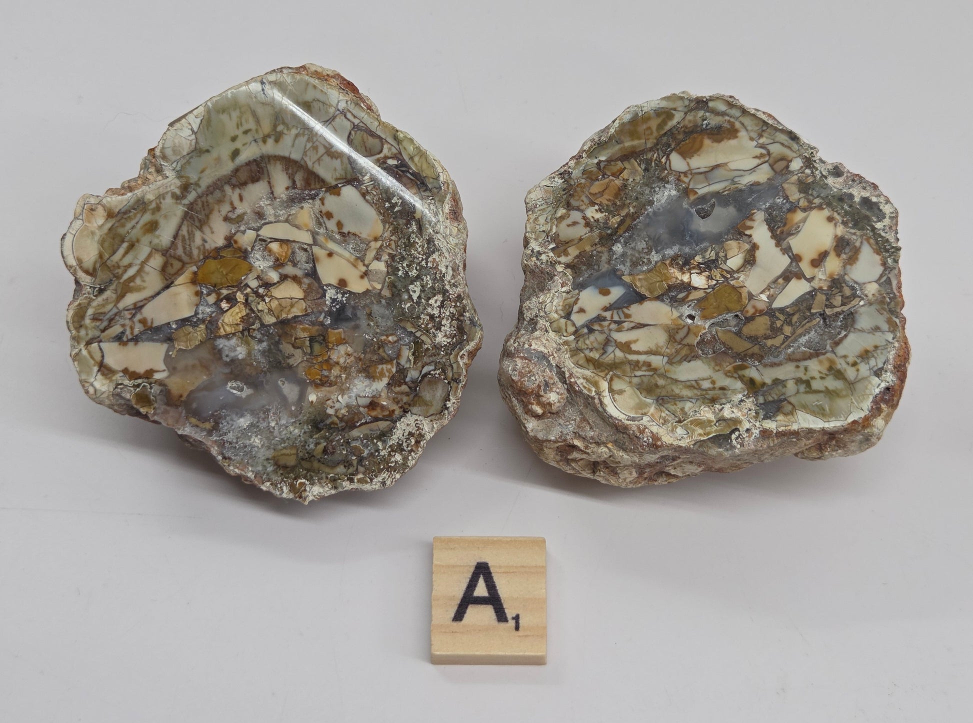 Two rough polished stone or mineral specimens with mixed beige, gray, brown, and metallic flecks, photographed on a light gray background. A small wooden letter tile with the letter A is placed to the left for size reference.