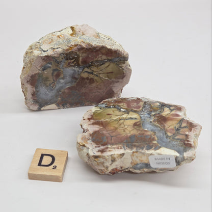 Two polished petrologically interesting raw stone slabs with a mix of brown, beige, gray, and blue-green minerals, displayed on a white background with a small wooden Scrabble tile showing the letter D2.