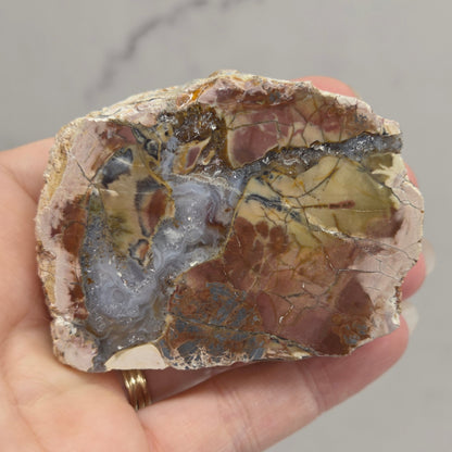 A hand holding a raw mineral specimen with a mix of brown, beige, gray, and metallic gold patterns, set against a light gray marble-patterned background.
