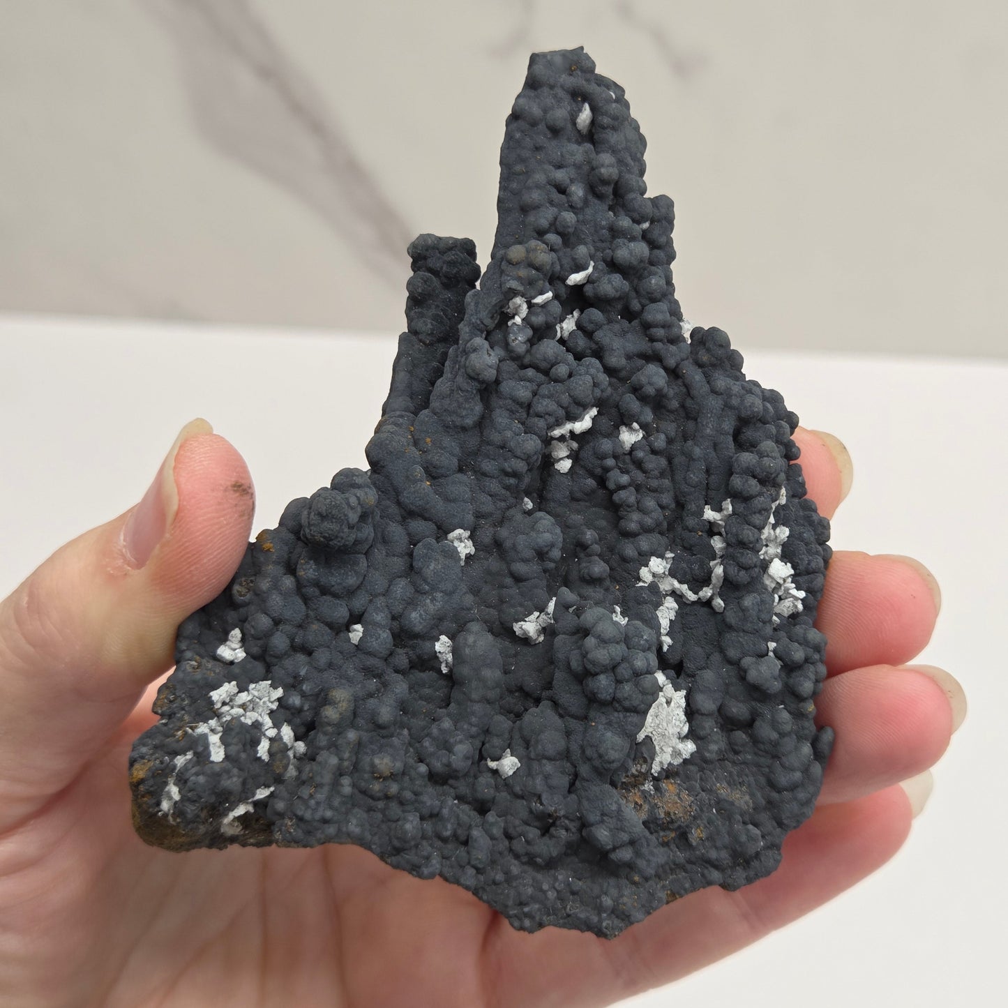 A hand holding a rough, porous dark gray mineral specimen with small white crystalline inclusions, photographed against a light indoor background.