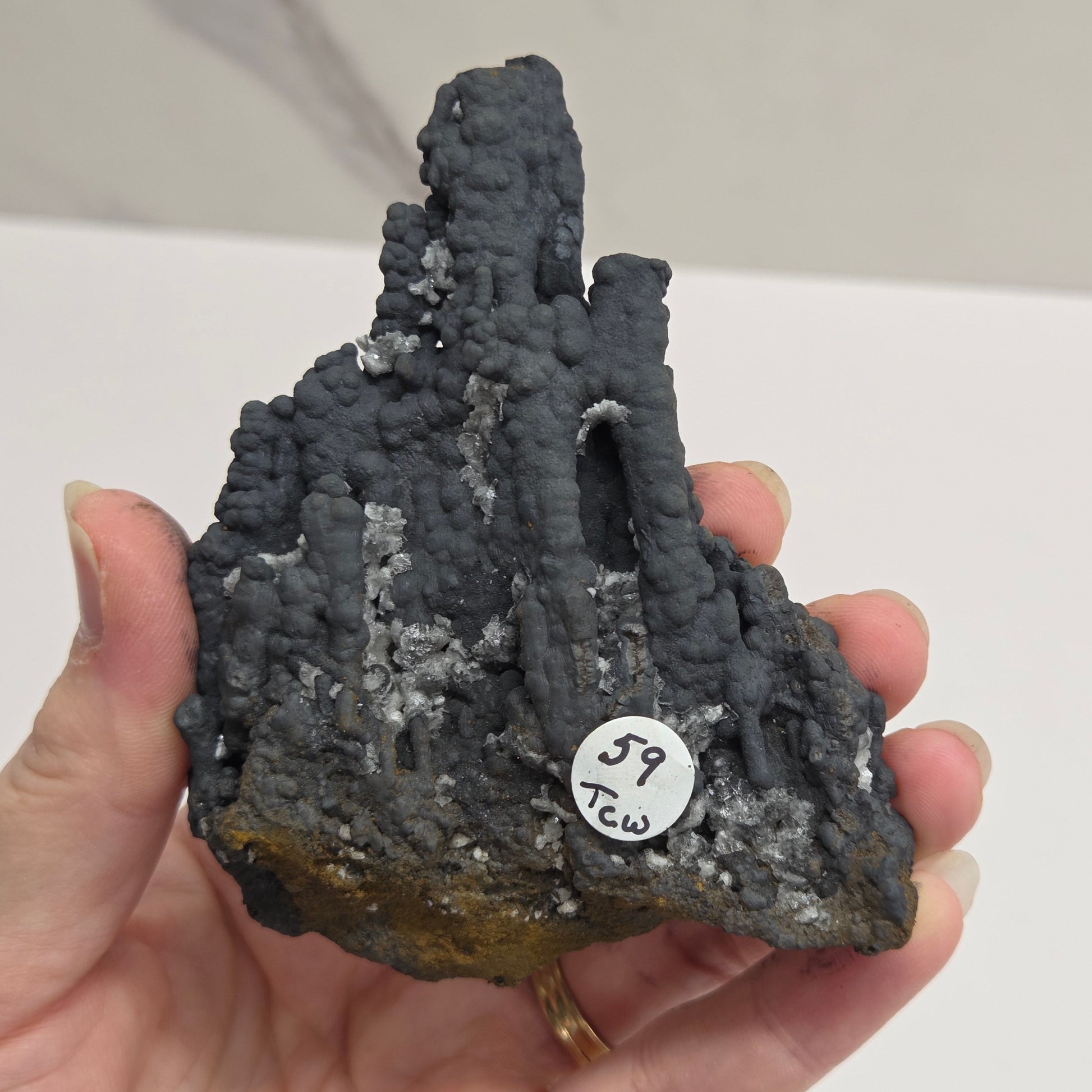 A hand holding a rough, porous dark gray mineral or rock specimen with a small white circular tag marked '57 K23'.