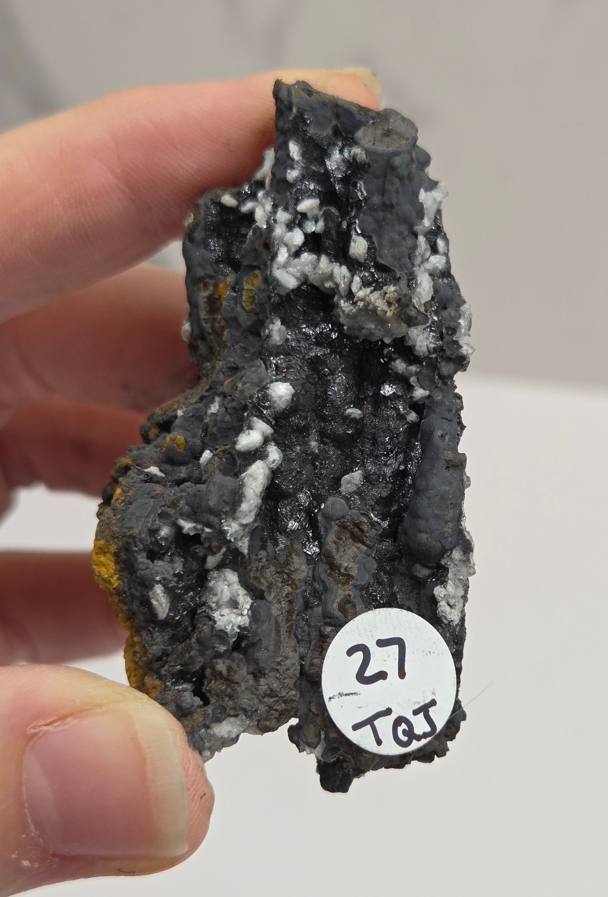 A hand holding a small rough mineral specimen with a white paper tag marked '27 101' against a light neutral background.