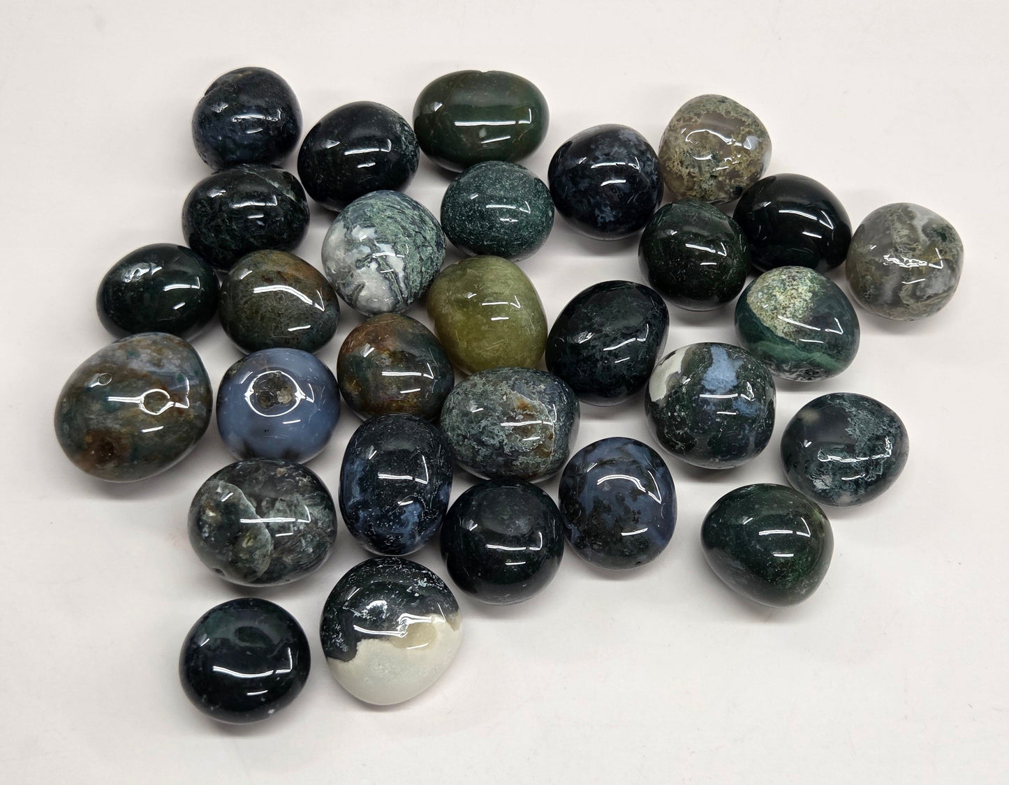 A pile of polished gemstone pendants or beads in various earthy and dark colors, including black, gray, green, and blue, arranged on a plain white background.