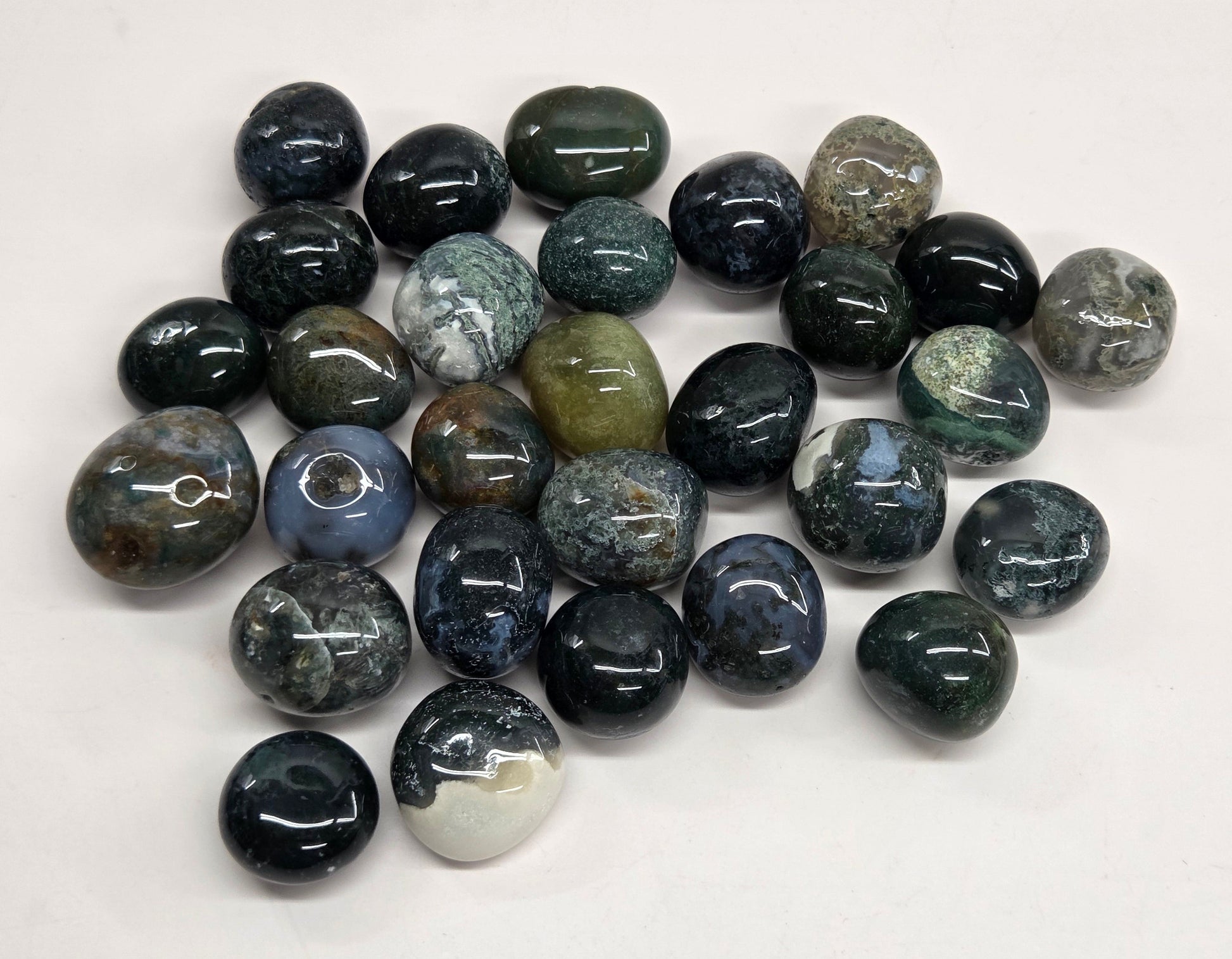A pile of polished gemstone pendants or beads in various earthy and dark colors, including black, gray, green, and blue, arranged on a plain white background.