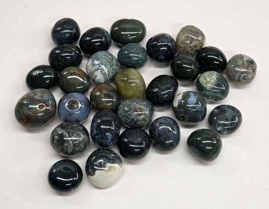 A pile of polished gemstone pendants or beads in various earthy and dark colors, including black, gray, green, and blue, arranged on a plain white background.