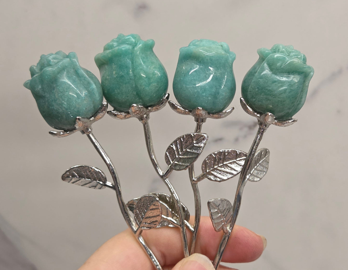 A hand holding four carved turquoise gemstone rose buds on silver metal stems with leaves, set against a light gray marble background.