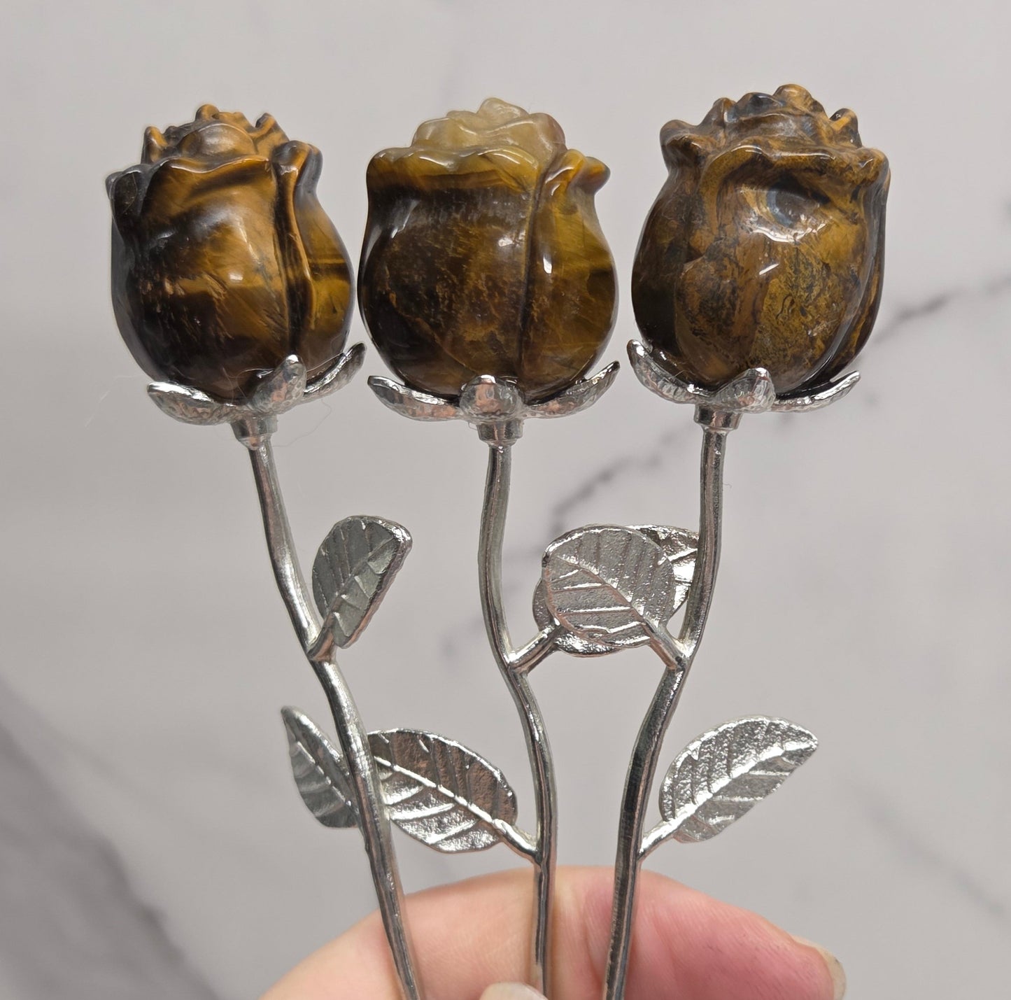 A close-up of a hand holding three carved brown rose-shaped objects on silver-toned stems with leaf details, set against a light marble-patterned background.