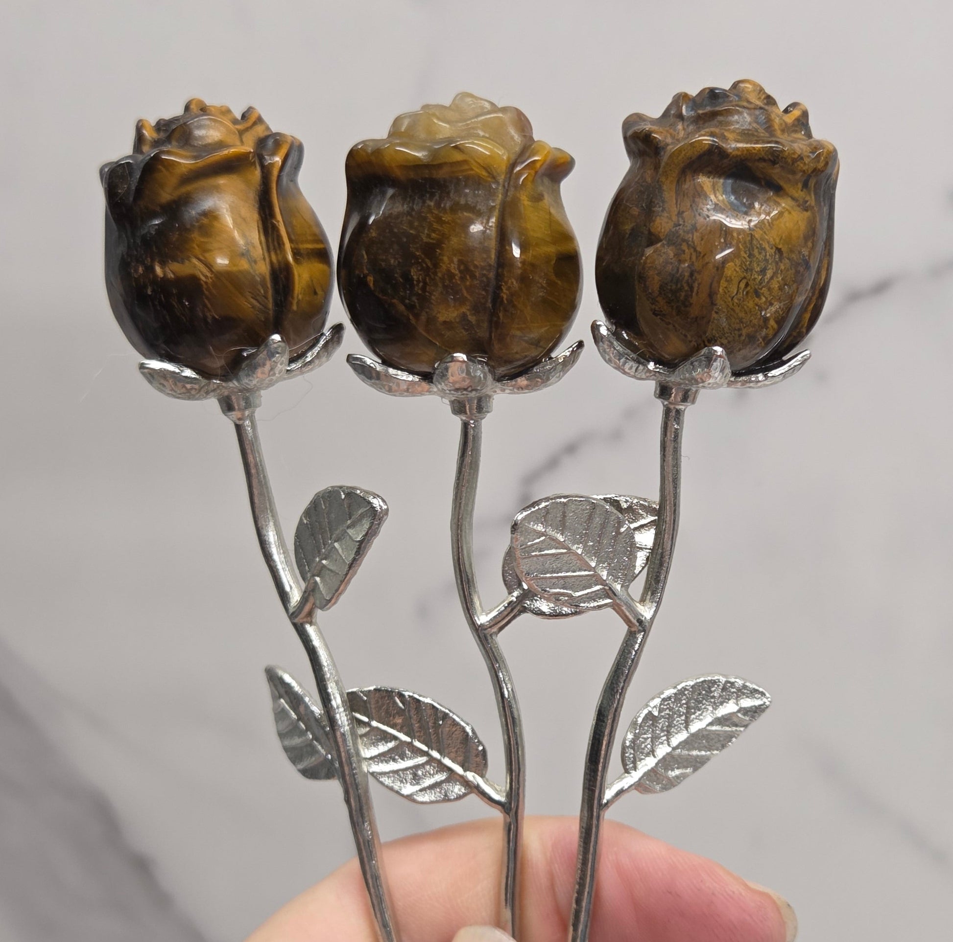 A close-up of a hand holding three carved brown rose-shaped objects on silver-toned stems with leaf details, set against a light marble-patterned background.