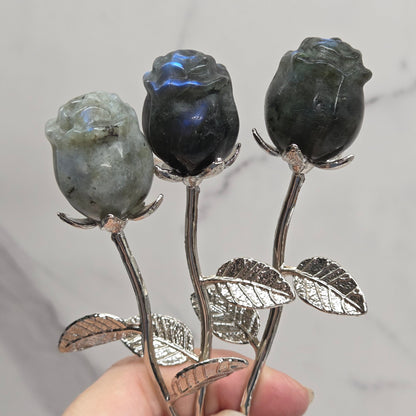 A close-up of a hand holding three lapel pins shaped like dark flowers with silver-toned stems and leaves, set against a light gray marble background.