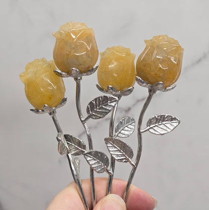 A hand holding five small rose-shaped crystal carvings with yellow translucent petals and silver metal stems and leaves, set against a light marble background.