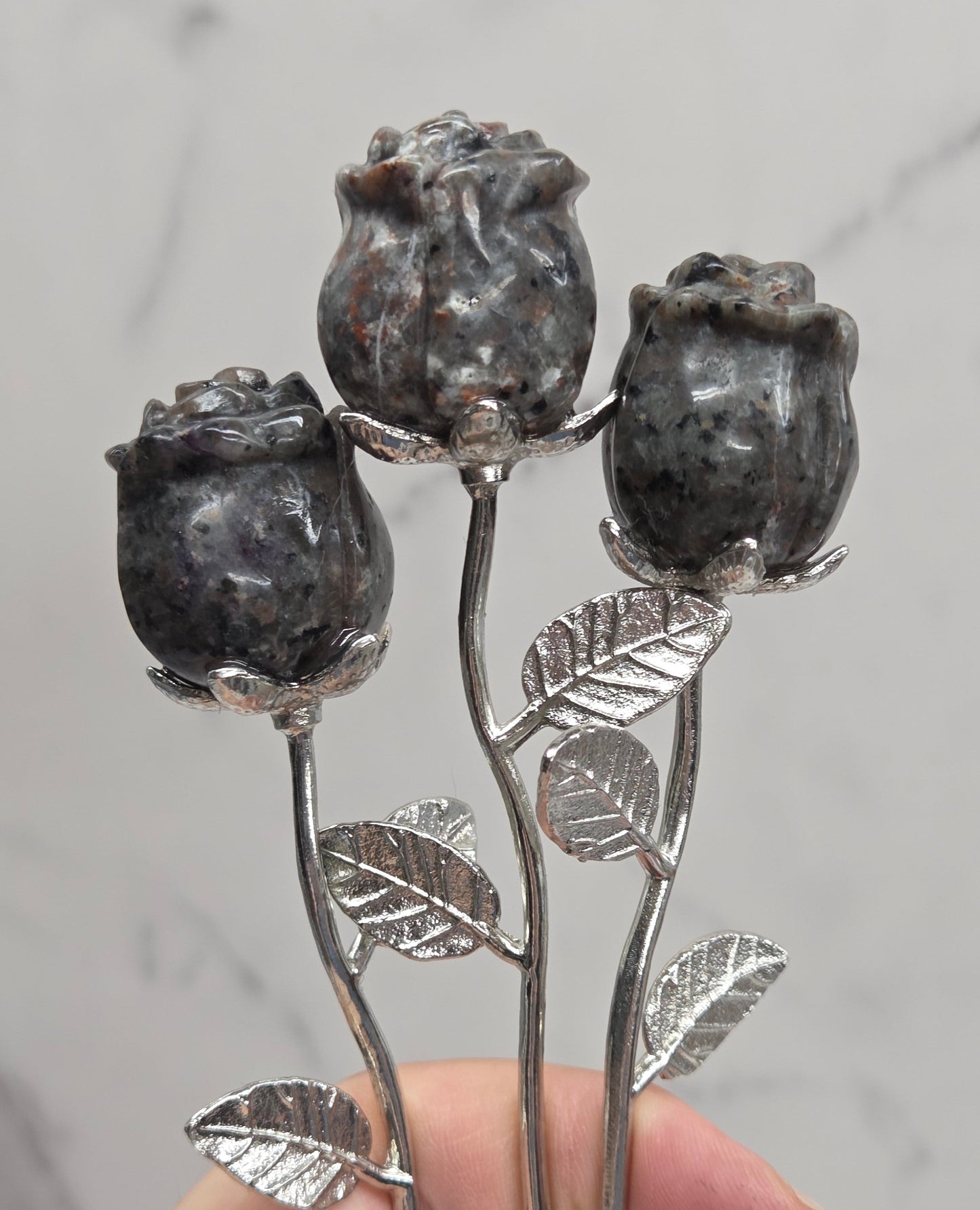 Three dark gray stone rose stems with silver-toned metal leaves and buds, held in a person's hand against a light gray marble background.