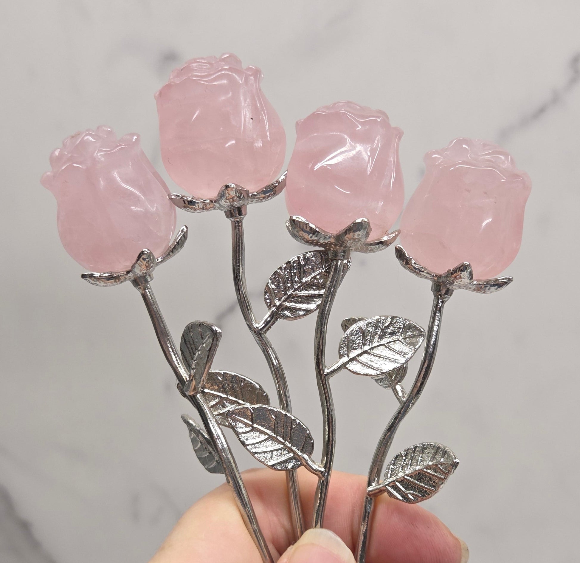 A hand holding a decorative rose-shaped carving with pink translucent rose heads and silver-toned stems and leaves, set against a light marble-patterned background.