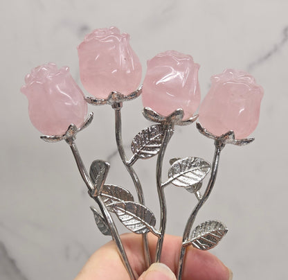 A hand holding a decorative rose-shaped carving with pink translucent rose heads and silver-toned stems and leaves, set against a light marble-patterned background.
