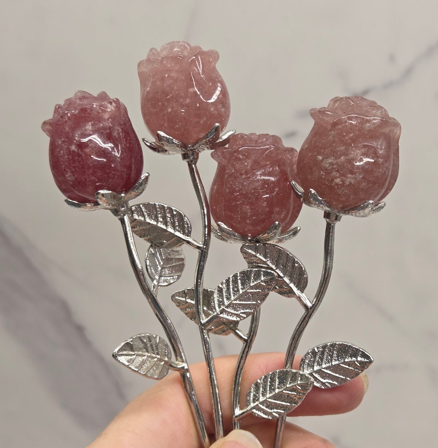A hand holding a silver-toned metal rose-shaped hair claw clip with four pinkish-red stone rose buds and detailed silver leaves, set against a light gray marble background.