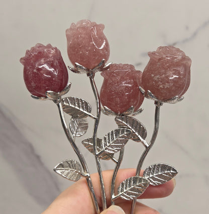 A hand holding a silver-toned metal rose-shaped hair claw clip with four pinkish-red stone rose buds and detailed silver leaves, set against a light gray marble background.