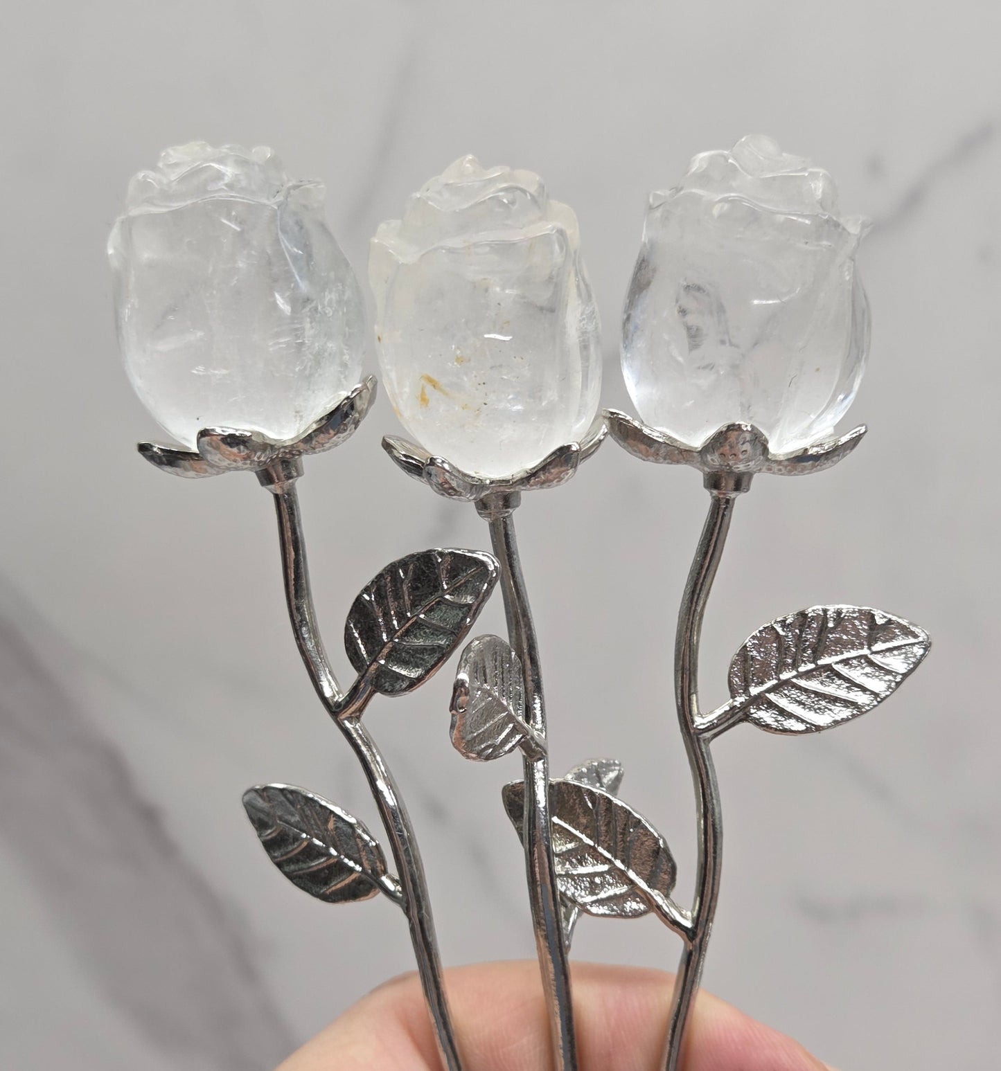 A hand holding three rose-shaped metal and crystal flower stems against a light gray marble background.