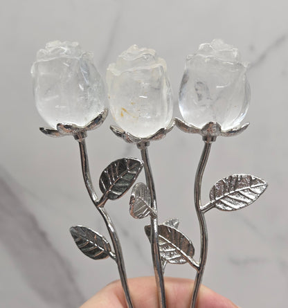 A hand holding three rose-shaped metal and crystal flower stems against a light gray marble background.