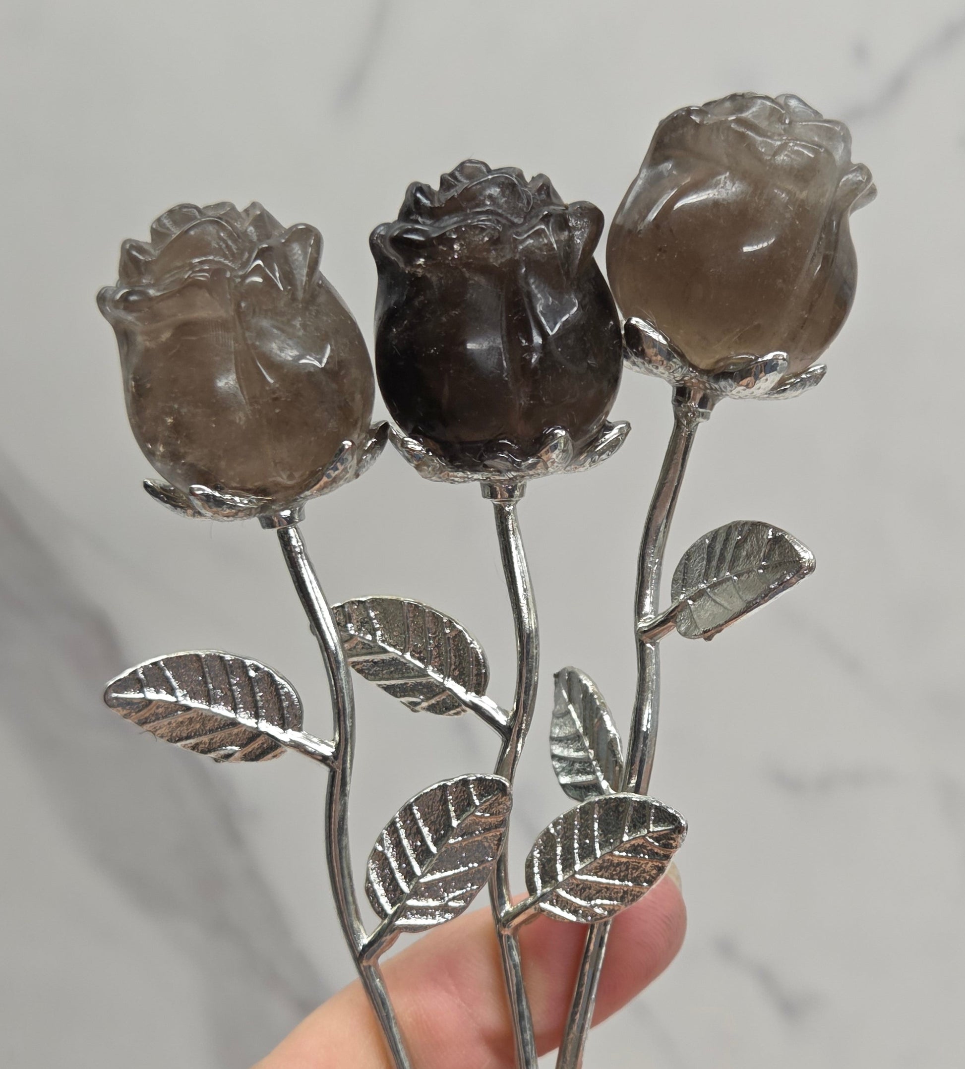 A hand holding three rose-shaped hairpins with carved smoky brown gemstone petals and silver-toned stems and leaves, set against a light gray marble background.