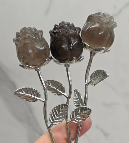 A hand holding three rose-shaped hairpins with carved smoky brown gemstone petals and silver-toned stems and leaves, set against a light gray marble background.