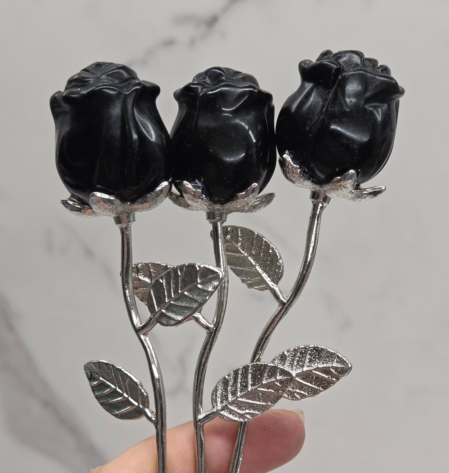 A hand holding three black rose buds on silver metal stems with textured leaves, set against a light gray marble-patterned background.