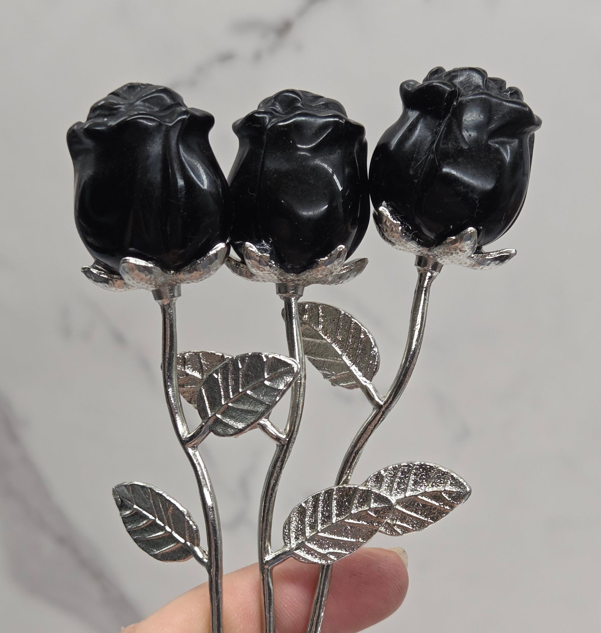 A hand holding three black rose buds on silver metal stems with textured leaves, set against a light gray marble-patterned background.