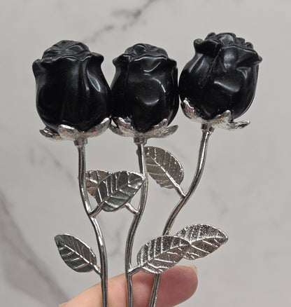 A hand holding three black rose buds on silver metal stems with textured leaves, set against a light gray marble-patterned background.