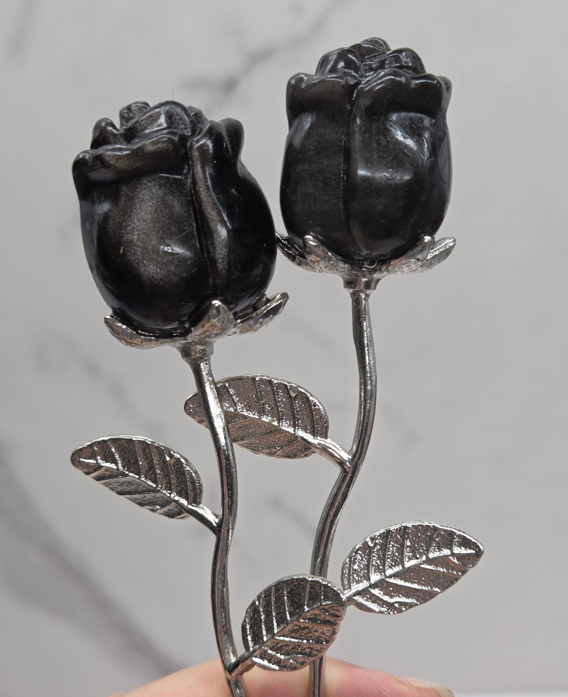 A close-up of a hand holding a silver-toned metal rose branch with two dark, polished rosebud charms against a light gray marble background.