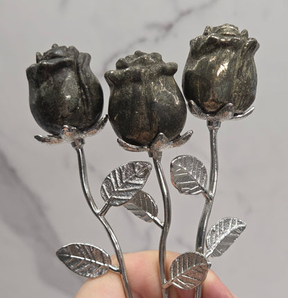 A hand holding three dark metallic rose-shaped hairpins with silver-toned stems and leaves against a light gray marble-patterned background.