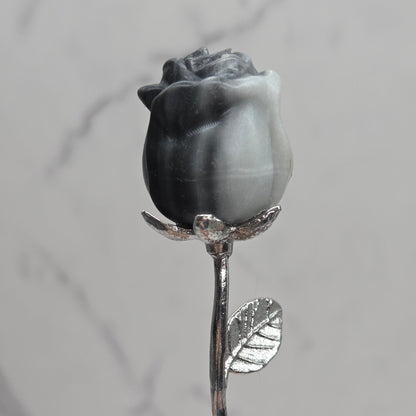 A hand holding a decorative silver-toned rose stem with a dark gray marbled rosebud against a light gray marble-patterned background.