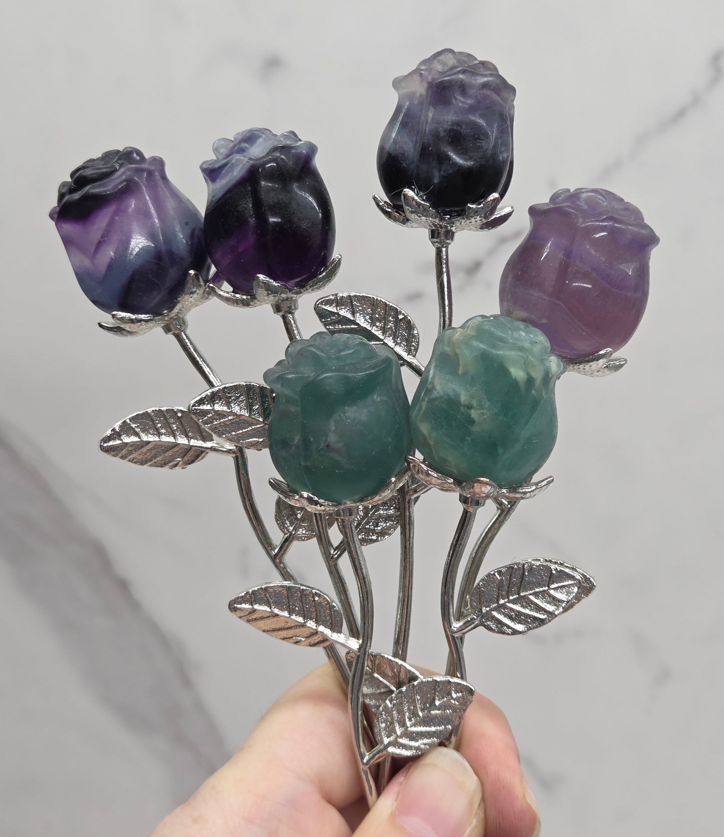 A hand holding a silver-toned floral brooch with carved purple and green gemstone rose buds and leaf details, set against a light gray marble background.