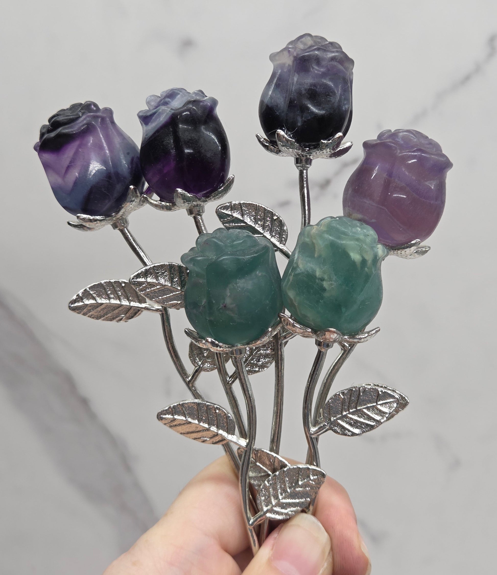 A hand holding a silver-toned floral brooch with carved purple and green gemstone rose buds and leaf details, set against a light gray marble background.